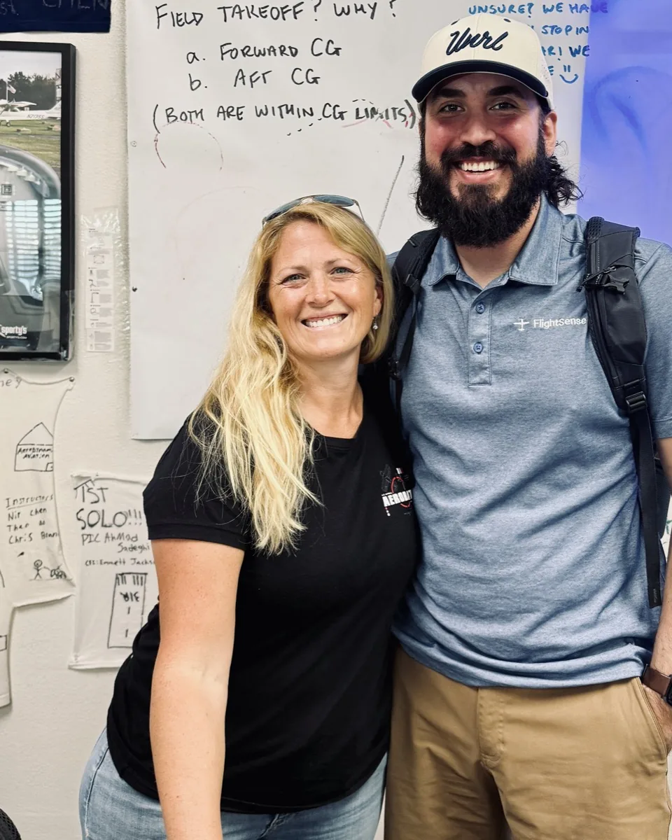 Jen Watson and Taylor Horsager standing together in the AeroDynamic Aviation briefing room, in front of a wall of first-solo shirts and a question-of-the-week whiteboard.